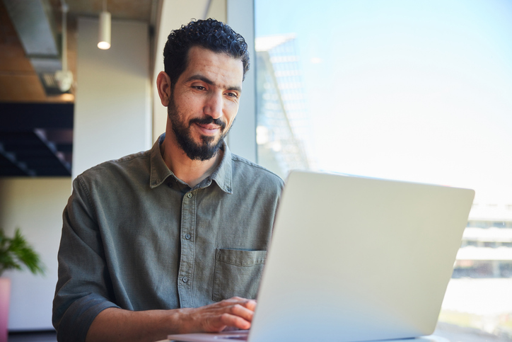 Smiling young businessman working on a laptop by an office window
