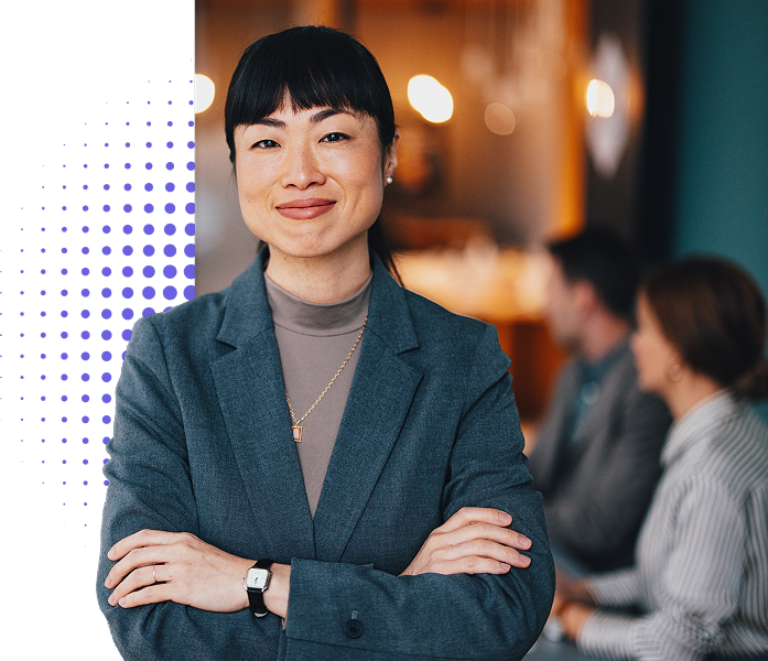 A person looks directly at the camera, crossing her arms and smiling while a group of people have a meeting in the background