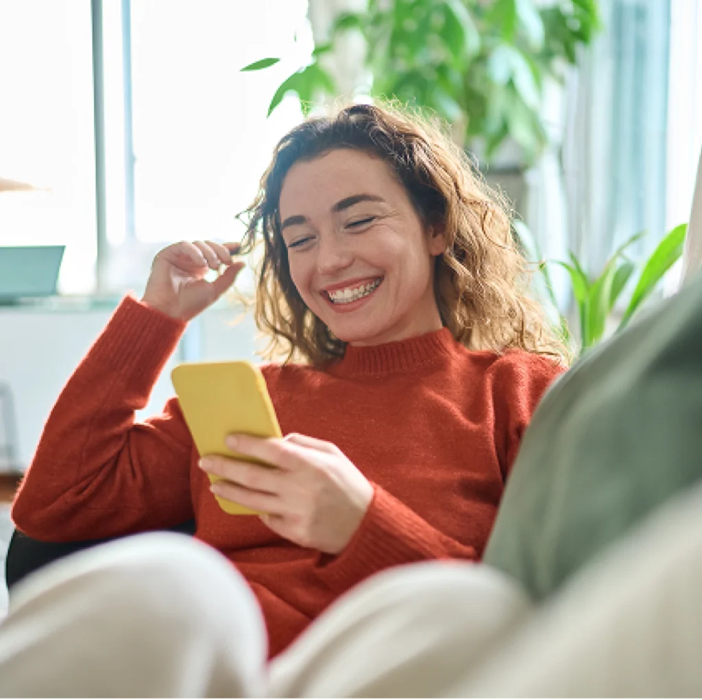 A woman smiles and laughs while relaxing on a couch looking at a phone