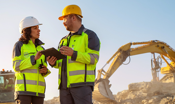 Two workers on an excavation site consult a tablet with heavy machinery in the background