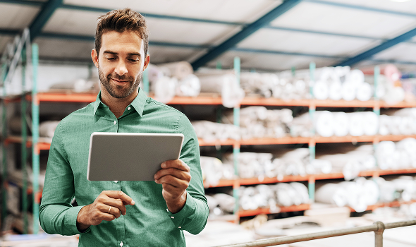 A person holds a tablet in front of a series of shelves stacked with rolls of an unknown product