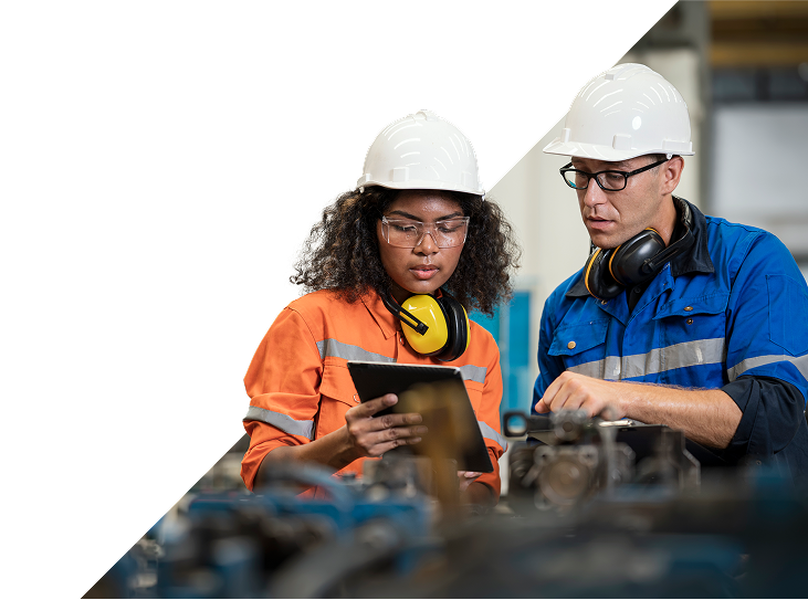 A man and a woman in safety jumpsuits, both wearing hard hats, safety glasses, and ear protection, review information on a tablet in an industrial setting.