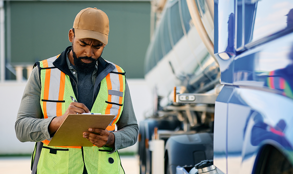 A worker writes on a clipboard while standing next to a tanker truck