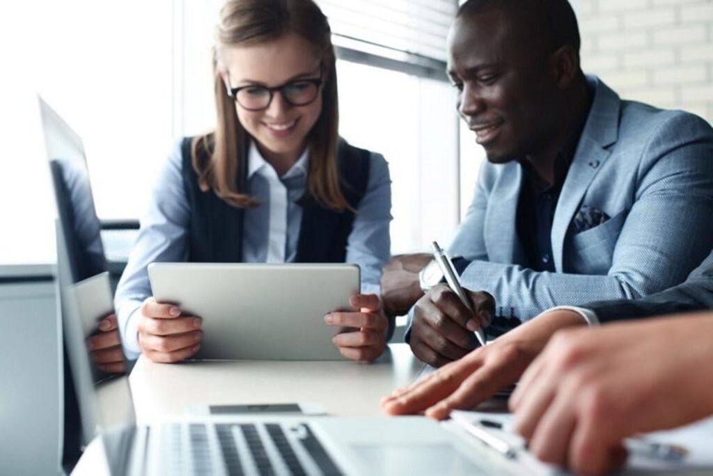 A group of people collaborate at a table, smiling and working on laptops and tablets