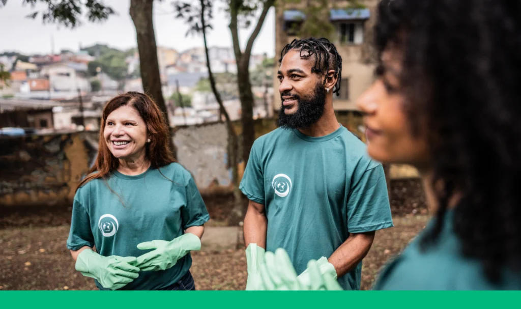 Three people wearing green shirts and rubber gloves stand outdoors, in a community area with trees and buildings in the background