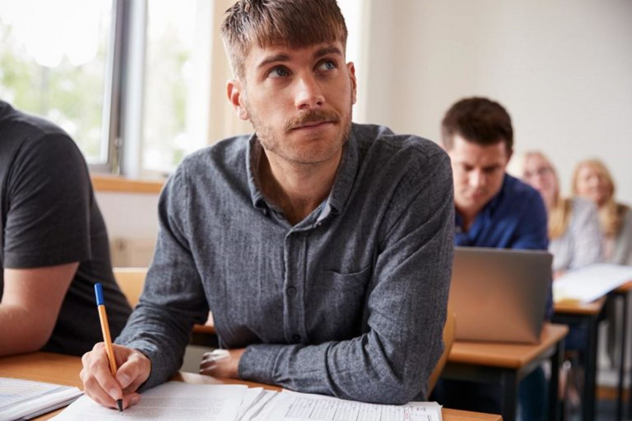 A man in a learning/training environment with others in the background, sits at a desk with a pen in his hand and a stack of papers in front of him