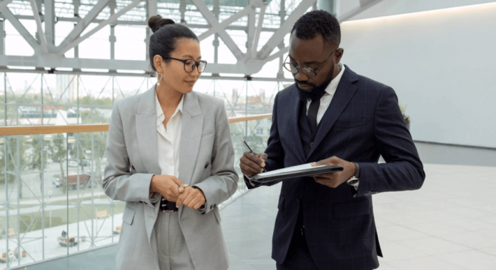 Man in suite writing on note pad while talking to woman in office building