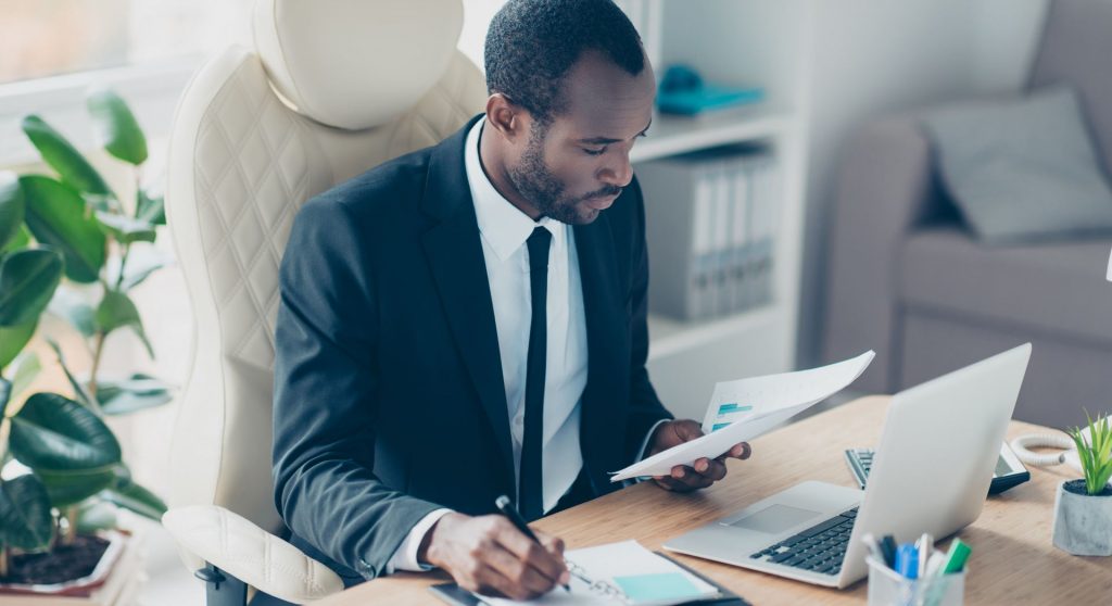 a man in an office at his desk looking at paper and a computer while writing down notes