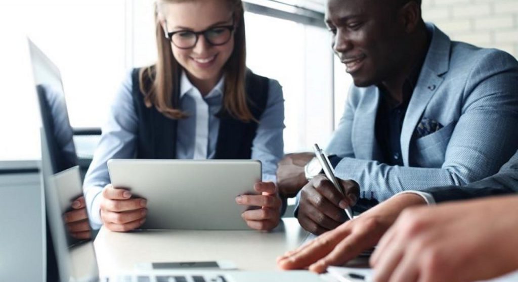A group of people collaborate at a table, smiling and working on laptops and tablets