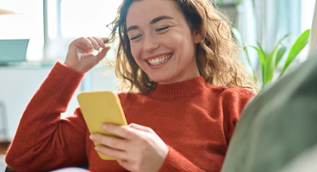 A woman smiles and laughs while relaxing on a couch looking at a phone