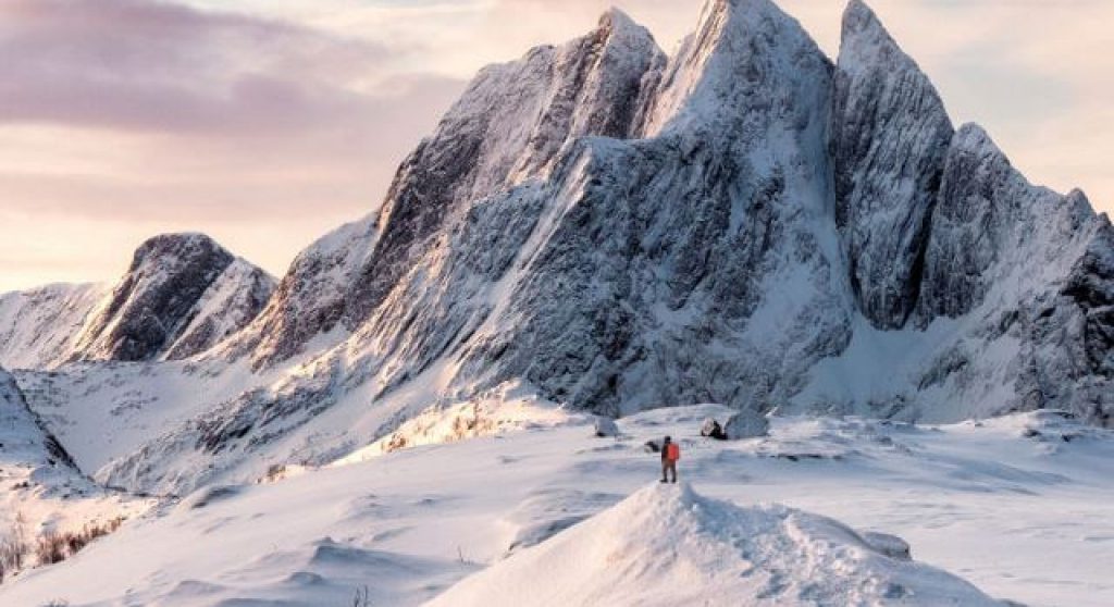 Man with backpack standing on snowy mountain peak during sunset