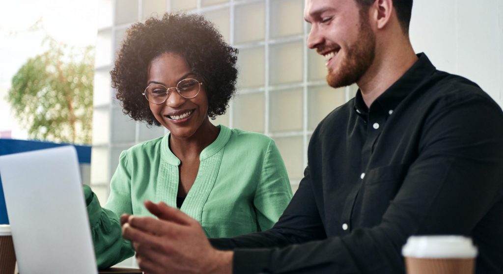 Two people sitting at a table outdoors, smiling at a laptop