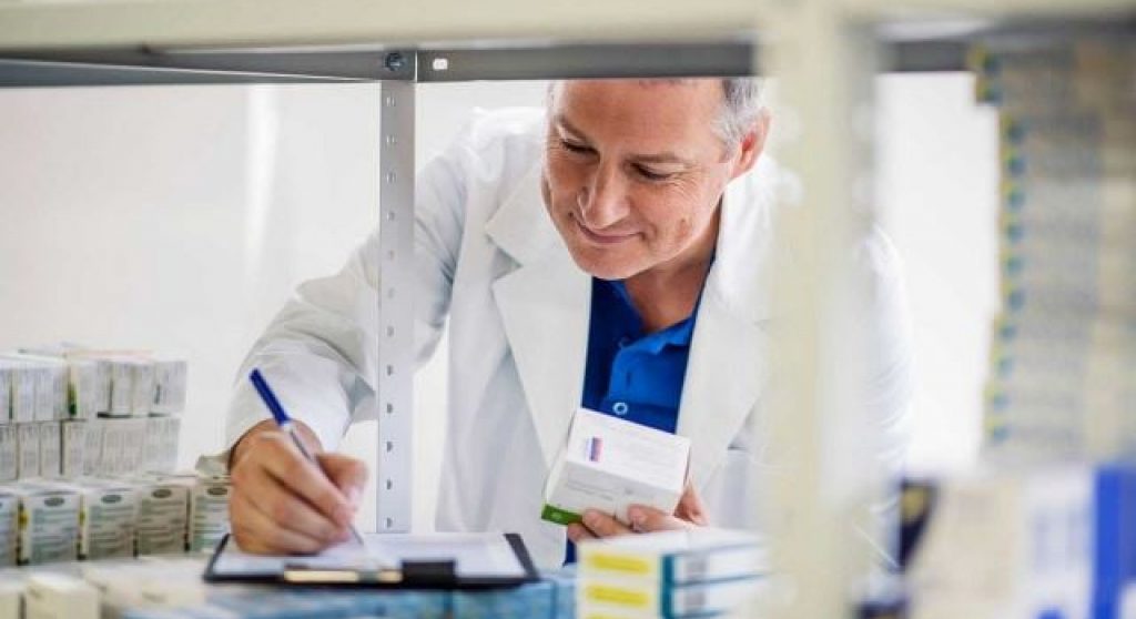 ein Apotheker schreibt Notizen auf einen Block, während er eine Bestandsaufnahme in einem Regal mit verschiedenen Medikamenten durchführt